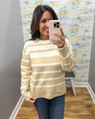 A woman takes a mirror selfie wearing a beige and white horizontally striped knit sweater and blue jeans in an indoor room with wood floors and a patterned wall.