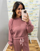 A woman takes a mirror selfie wearing a matching dusty rose sweatshirt and jogger pants set. The outfit features textured, ribbed knit fabric with unique draped details on the sleeves of the sweatshirt and a matching drawstring waist on the pants.