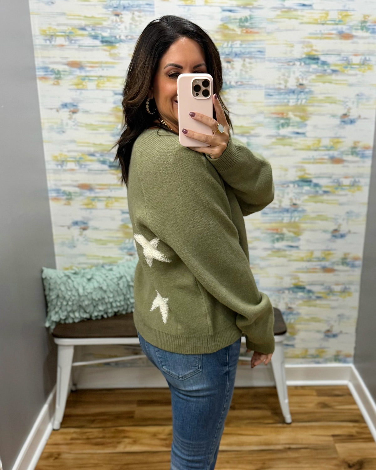 A woman takes a mirror selfie wearing an olive green knit sweater with white star patterns on the back, paired with blue jeans, in front of a patterned wallpaper background.