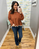 A woman takes a mirror selfie in a home hallway wearing a rust-orange long-sleeve knit sweater with a scalloped hem, blue jeans, and tan boots.