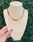 Chansuttpearls Herringbone gold snake chain necklace displayed on a beige fabric jewelry bust, held by a hand against a green leafy hedge background.