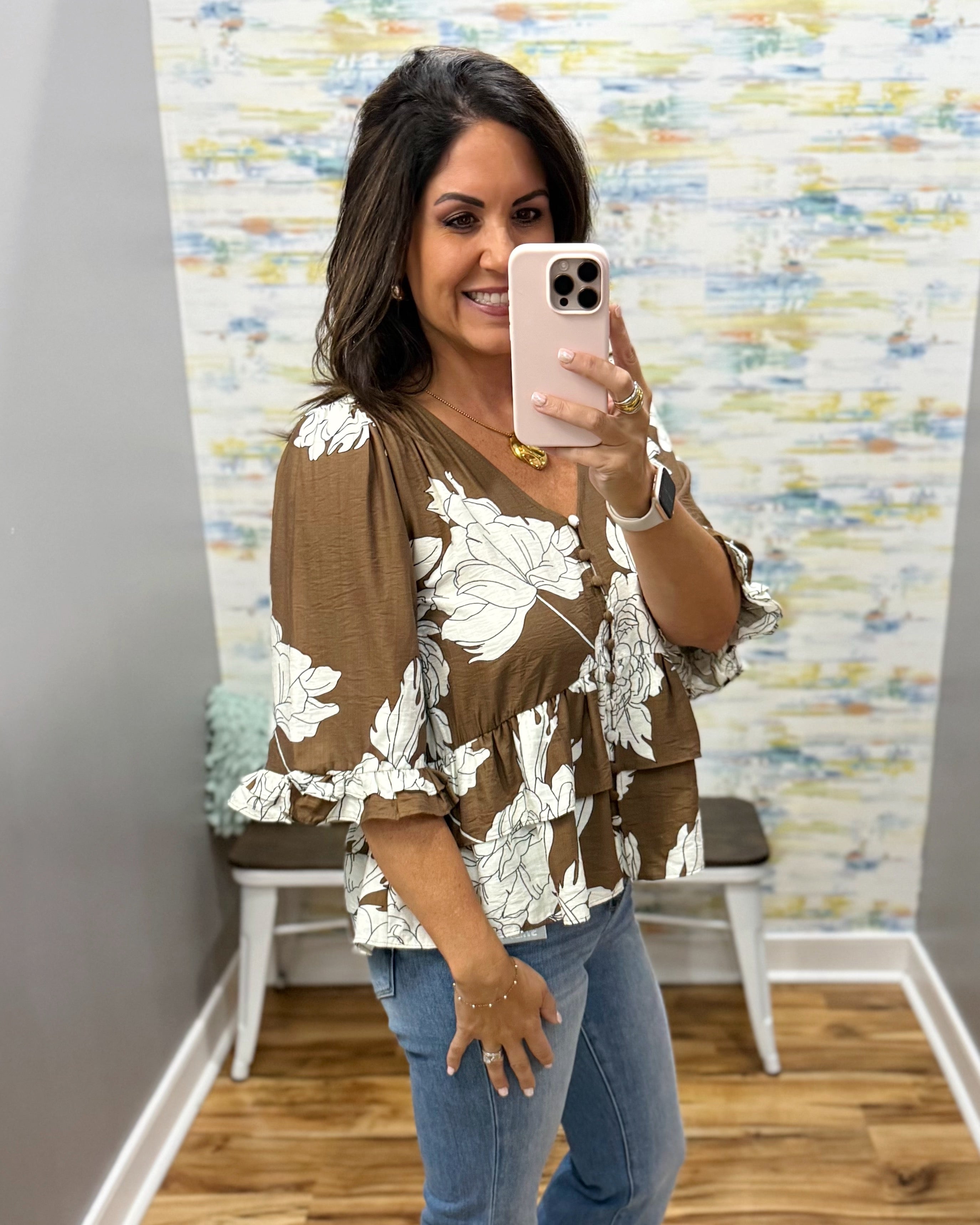 A woman takes a mirror selfie wearing a brown V-neck blouse with a white floral print and tiered ruffle sleeves, paired with blue jeans.