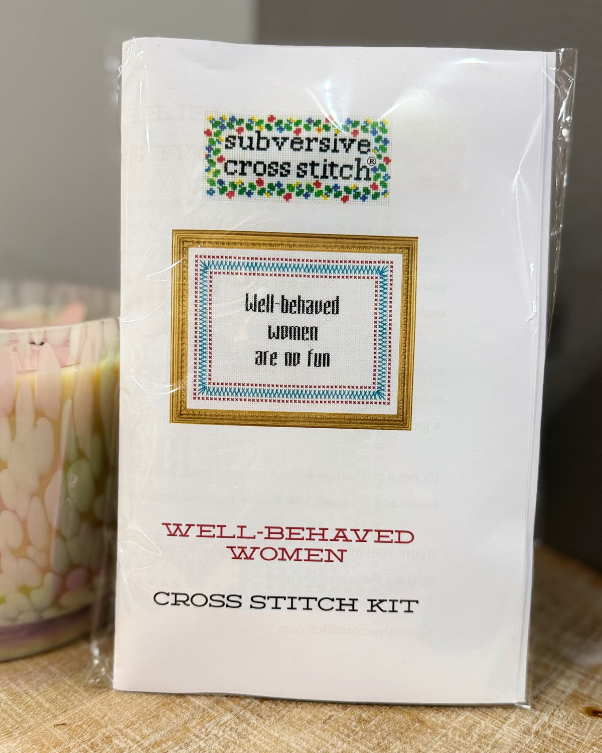 A packaged cross stitch kit labeled 'subversive cross stitch' and 'WELL-BEHAVED WOMEN CROSS STITCH KIT' on a white card, photographed in clear plastic wrap with a candle partially visible at the left edge.