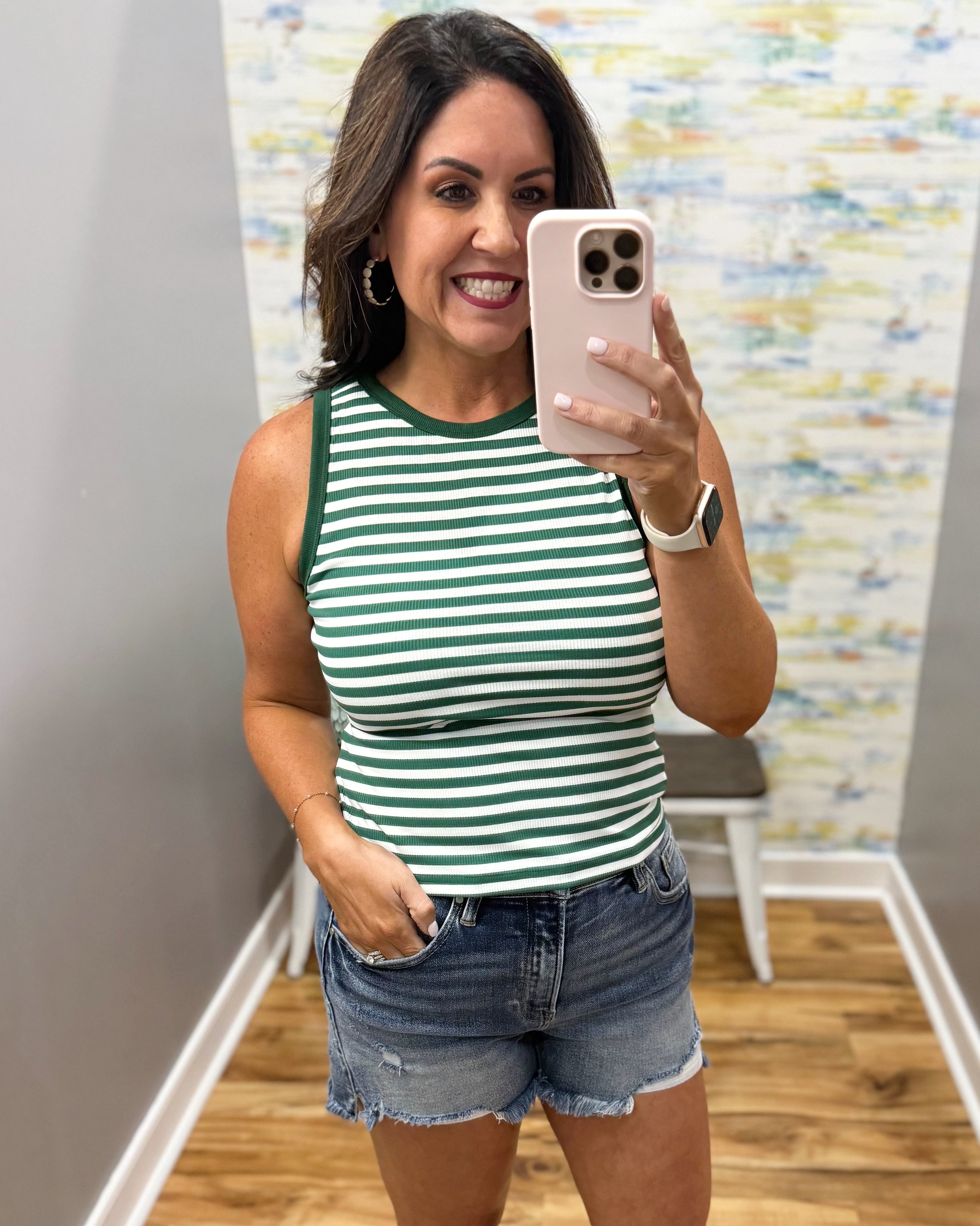 Woman taking a mirror selfie wearing a green and white striped tank top and denim shorts.