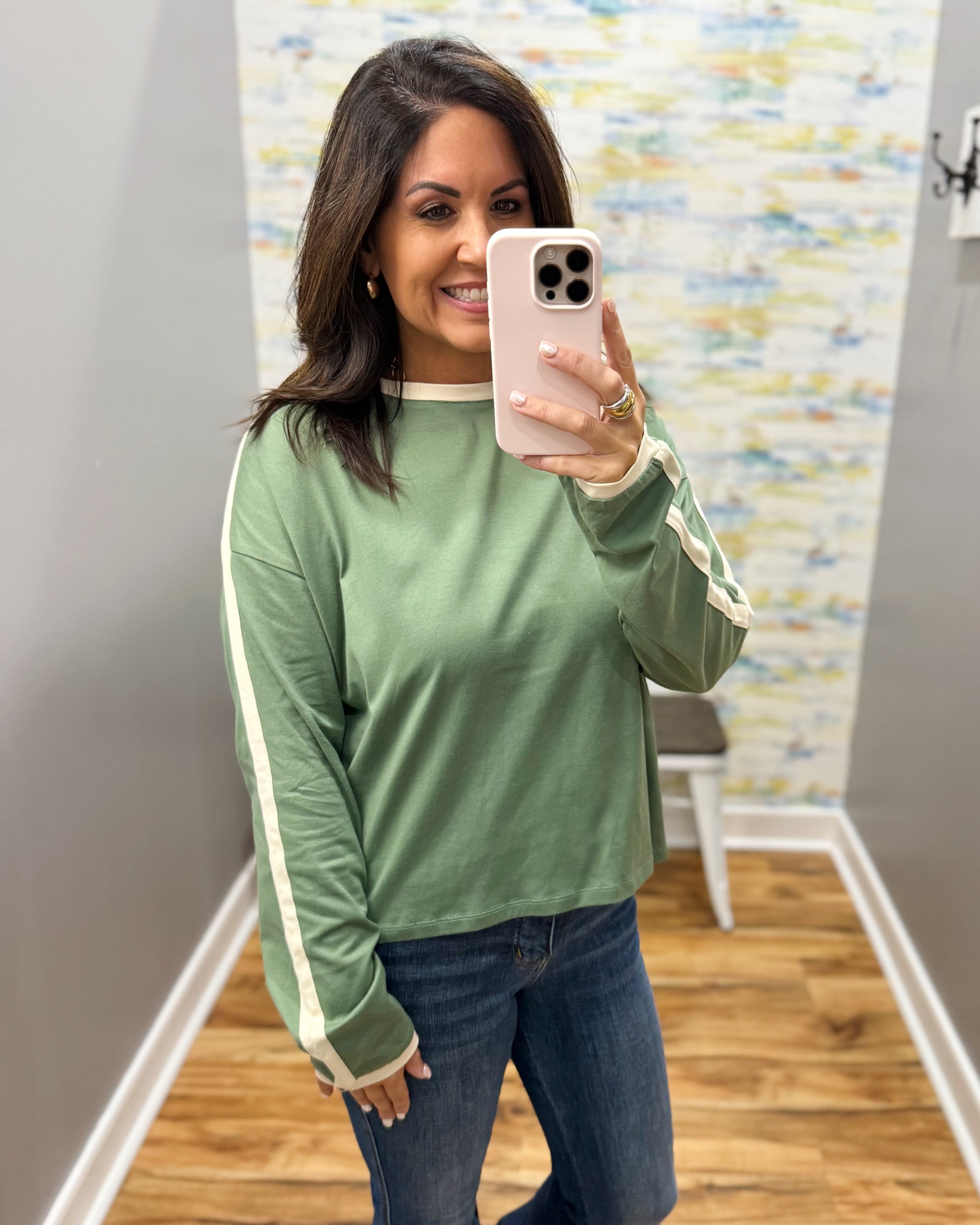 A woman takes a mirror selfie wearing a sage green long-sleeve crewneck shirt with cream stripes down the sleeves and cuffs, paired with blue jeans.