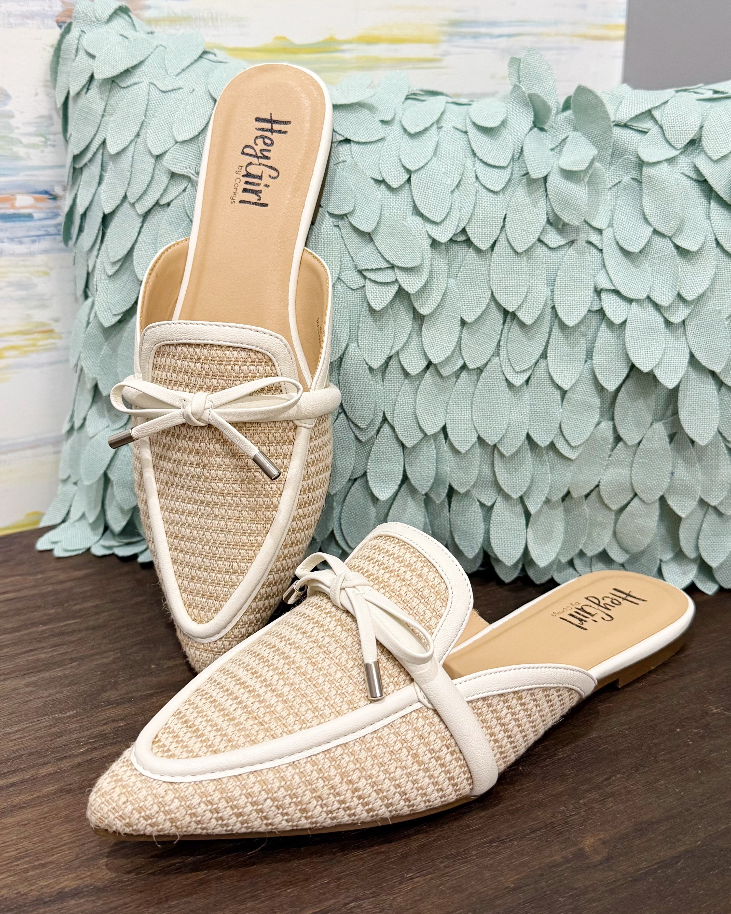 Pair of woven mules with white laces on a wooden surface with a decorative pillow in the background.