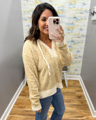 A woman takes a mirror selfie wearing a textured cream and gold knit sweater with a white drawstring, paired with blue jeans, in an indoor fitting room or home hallway.