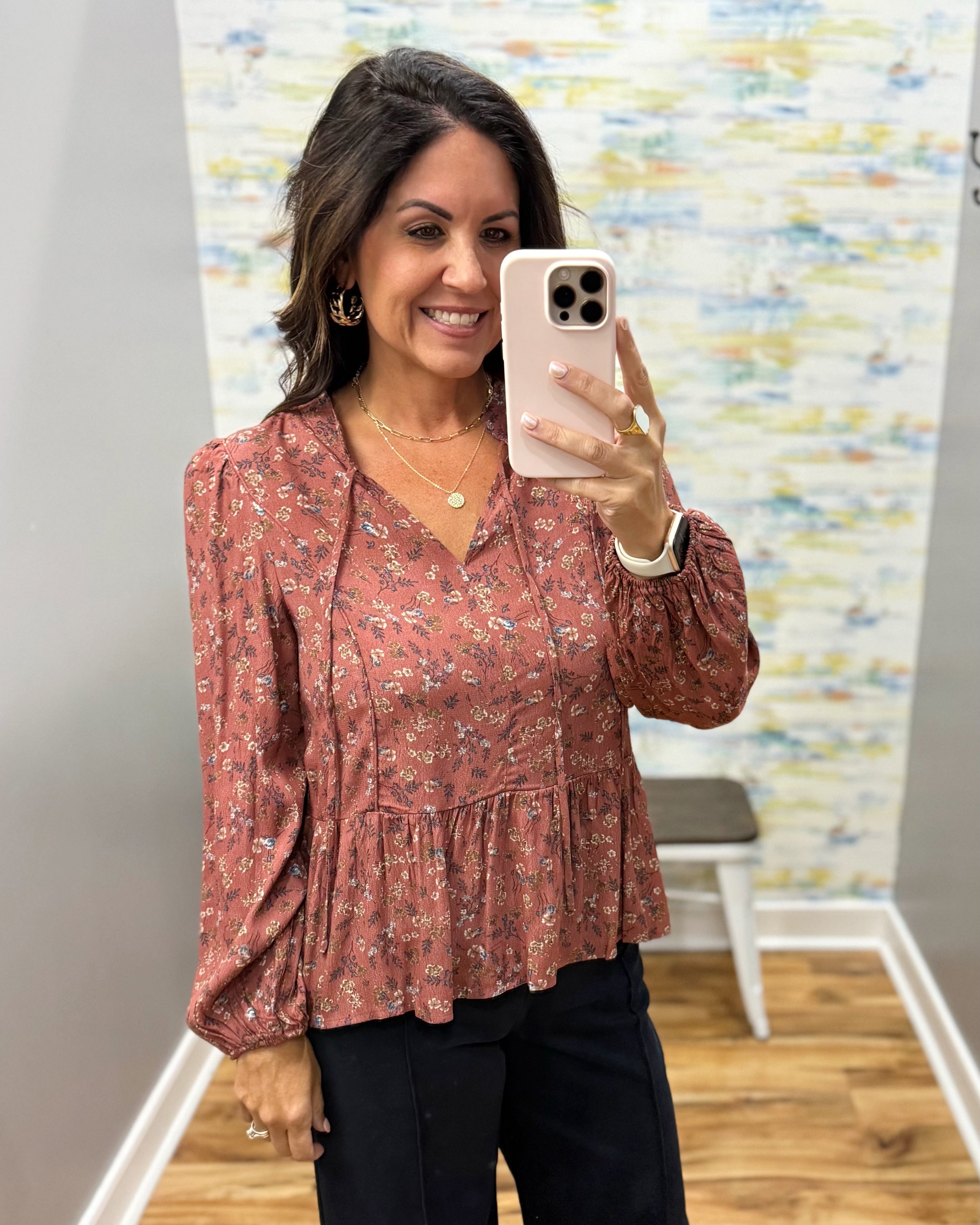 A woman takes a mirror selfie wearing a dusty rose long-sleeve blouse with a small white floral print and black pants in a fitting room.