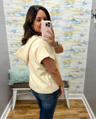 A woman takes a mirror selfie in a fitting room wearing a cream-colored knit hooded sweater vest and blue jeans.