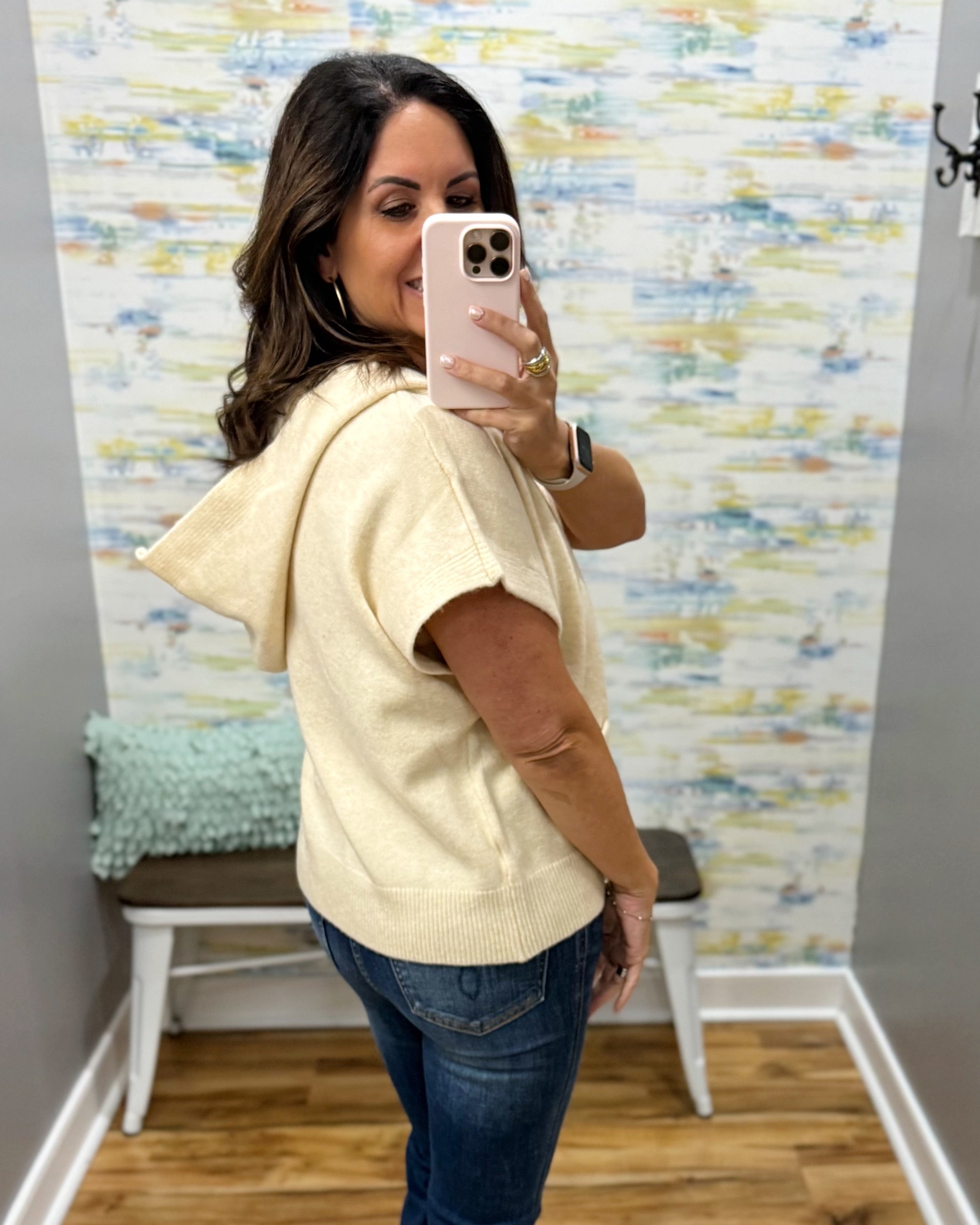 A woman takes a mirror selfie in a fitting room wearing a cream-colored knit hooded sweater vest and blue jeans.
