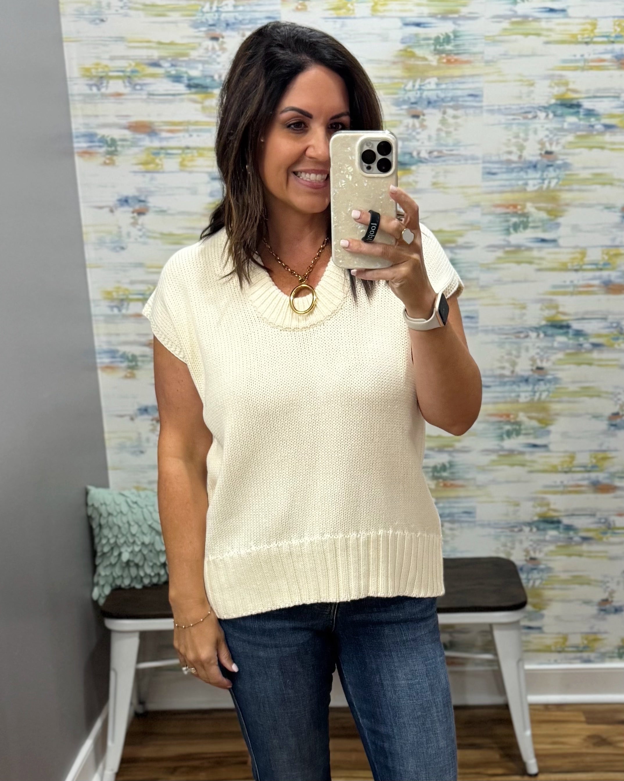 A woman takes a mirror selfie in a boutique showing a cream-colored short-sleeve knit sweater vest paired with dark blue jeans.