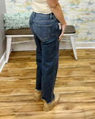 A person wearing blue Risen crop straight-leg jeans with a raw frayed hem, paired with beige boots and a cream top, standing on a wood floor in front of a white bench.