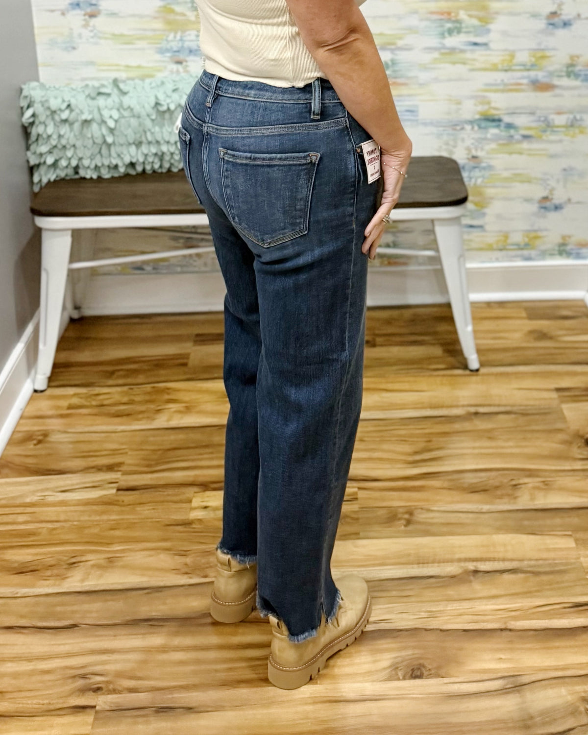 A person wearing blue Risen crop straight-leg jeans with a raw frayed hem, paired with beige boots and a cream top, standing on a wood floor in front of a white bench.