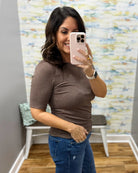 A woman taking a mirror selfie in a fitting room wearing a fitted short-sleeve brown top and blue jeans.