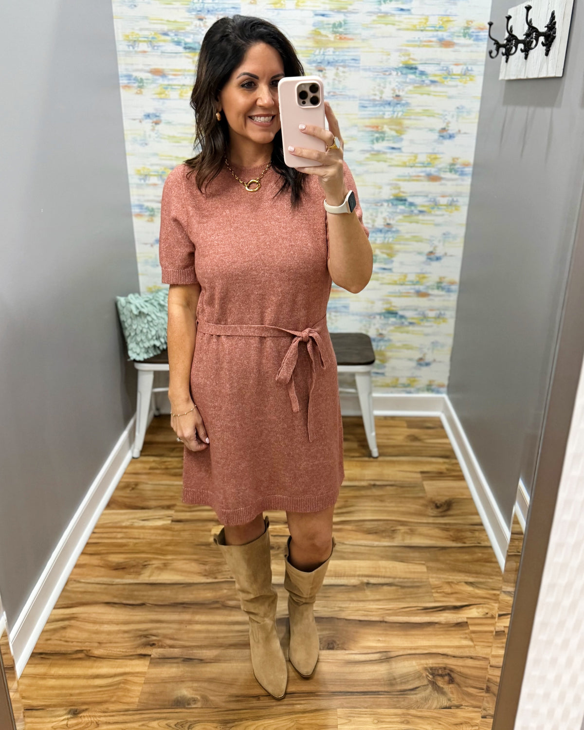 A woman takes a mirror selfie wearing a dusty rose short-sleeve tie-front dress and beige knee-high boots in a boutique fitting room with wood floors and a patterned wall.