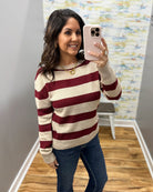 A woman takes a mirror selfie wearing a cream and burgundy striped knit sweater, blue jeans, and a gold necklace in an indoor room with a wood floor and abstract wall art.