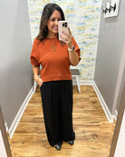 A woman takes a mirror selfie in a fitting room wearing a rust-orange short-sleeve knit top and black wide-leg pants.