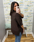 A woman takes a mirror selfie in a fitting room wearing a dark brown short-sleeve top and blue jeans.