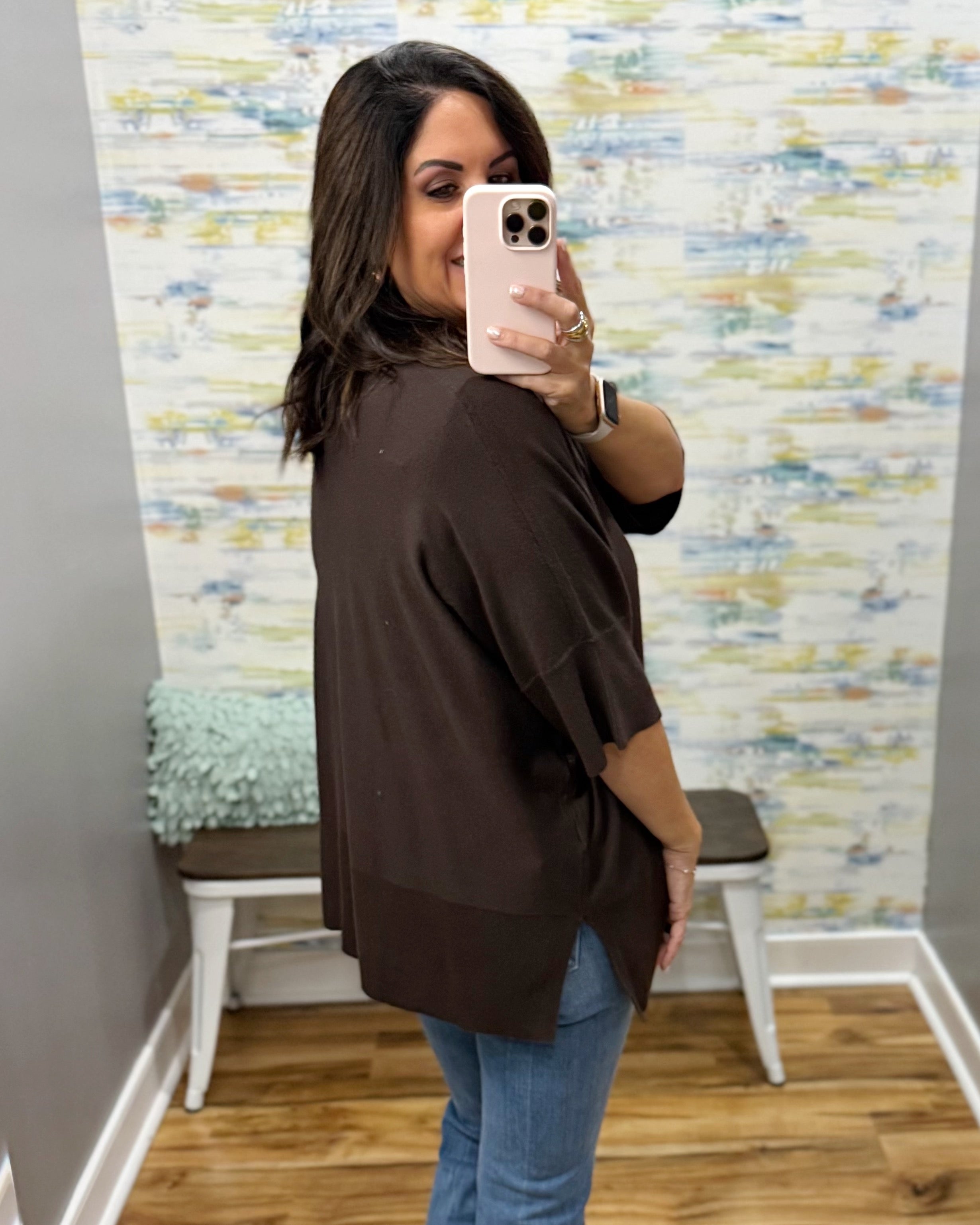 A woman takes a mirror selfie in a fitting room wearing a dark brown short-sleeve top and blue jeans.