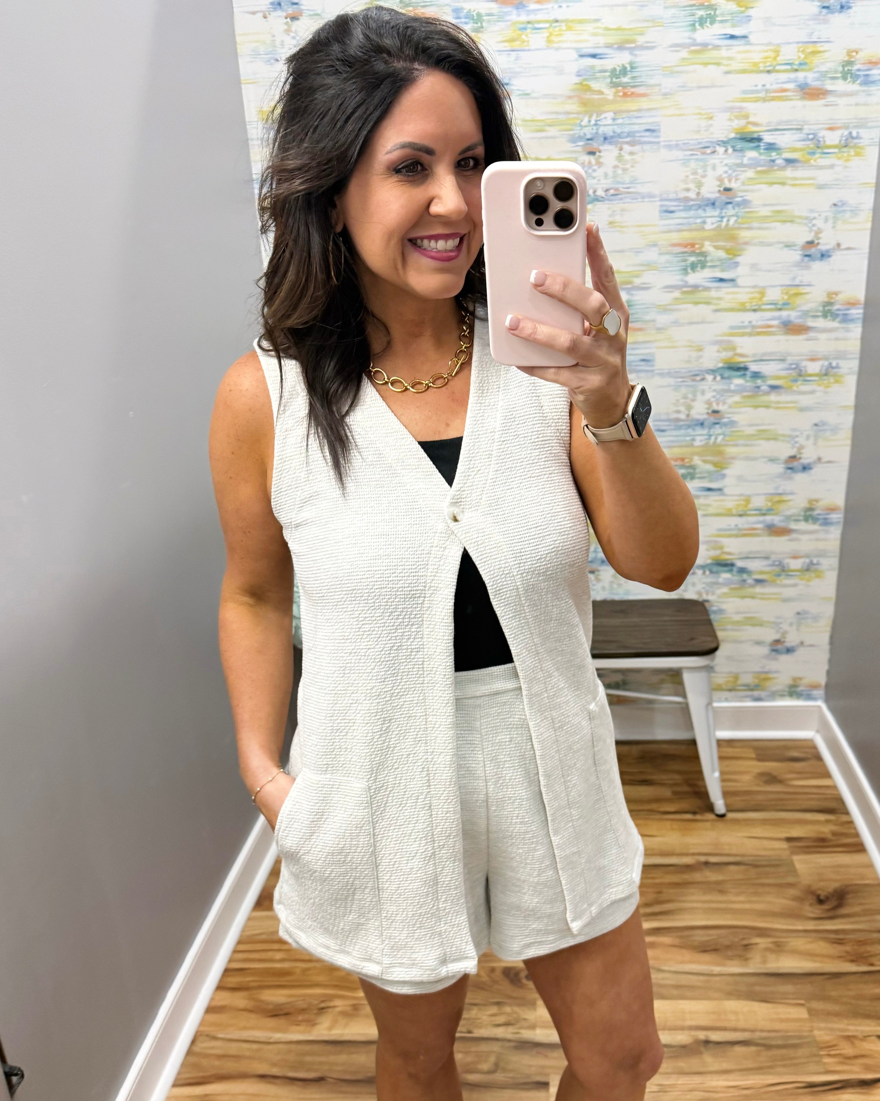 Woman taking a mirror selfie wearing a white vest and shorts in a room with a textured wall.