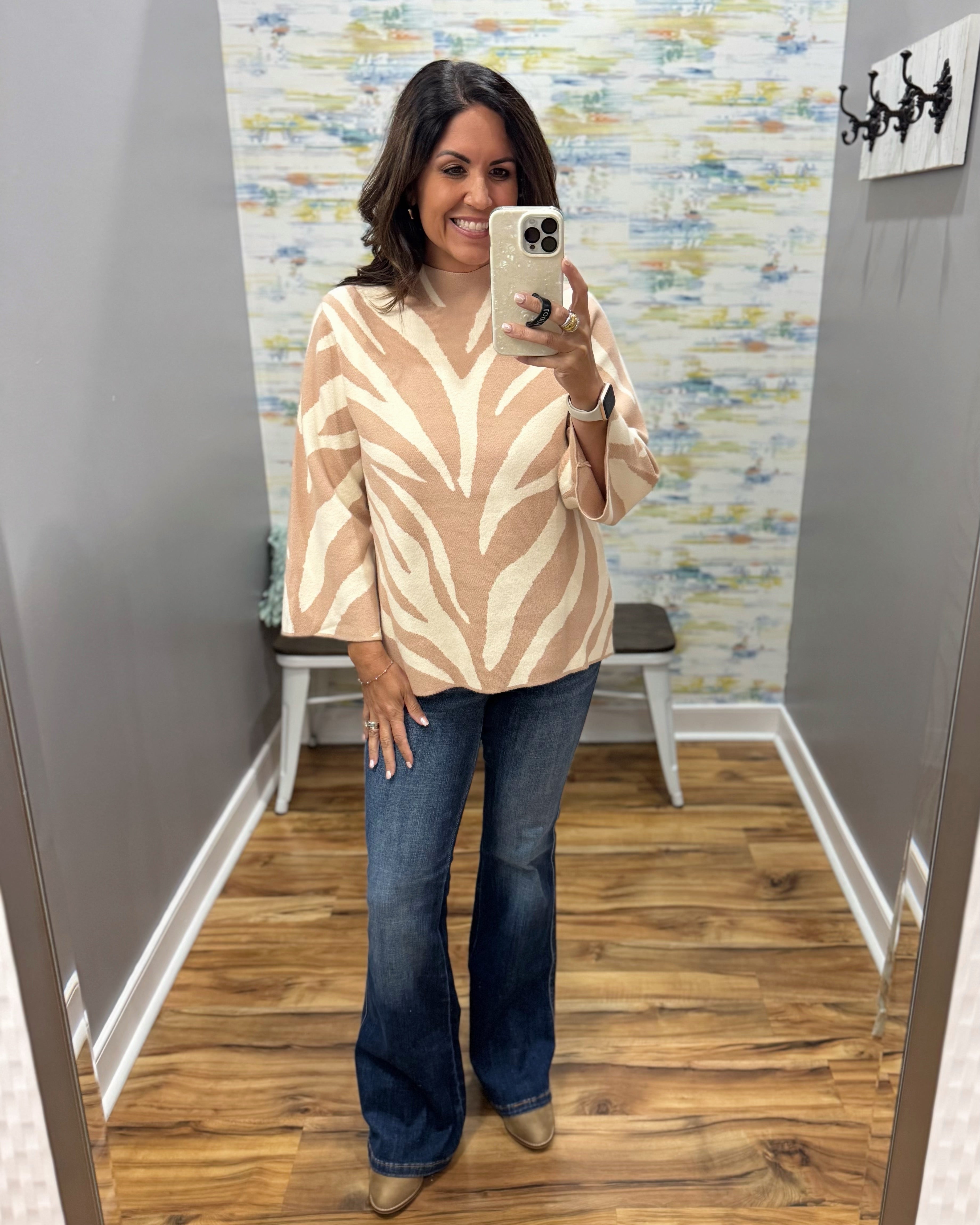 A woman takes a mirror selfie wearing a beige and cream wavy patterned knit top, blue flare jeans, and tan boots in a fitting room.