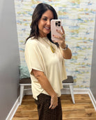 A woman taking a mirror selfie in a fitting room wearing a cream-colored short-sleeve top and brown leopard-print pants. She is holding a light pink smartphone.