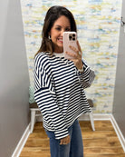 A woman takes a mirror selfie wearing a navy and white horizontally striped long-sleeve top, blue jeans, and a white undershirt in a fitting room with a patterned wallpaper background.