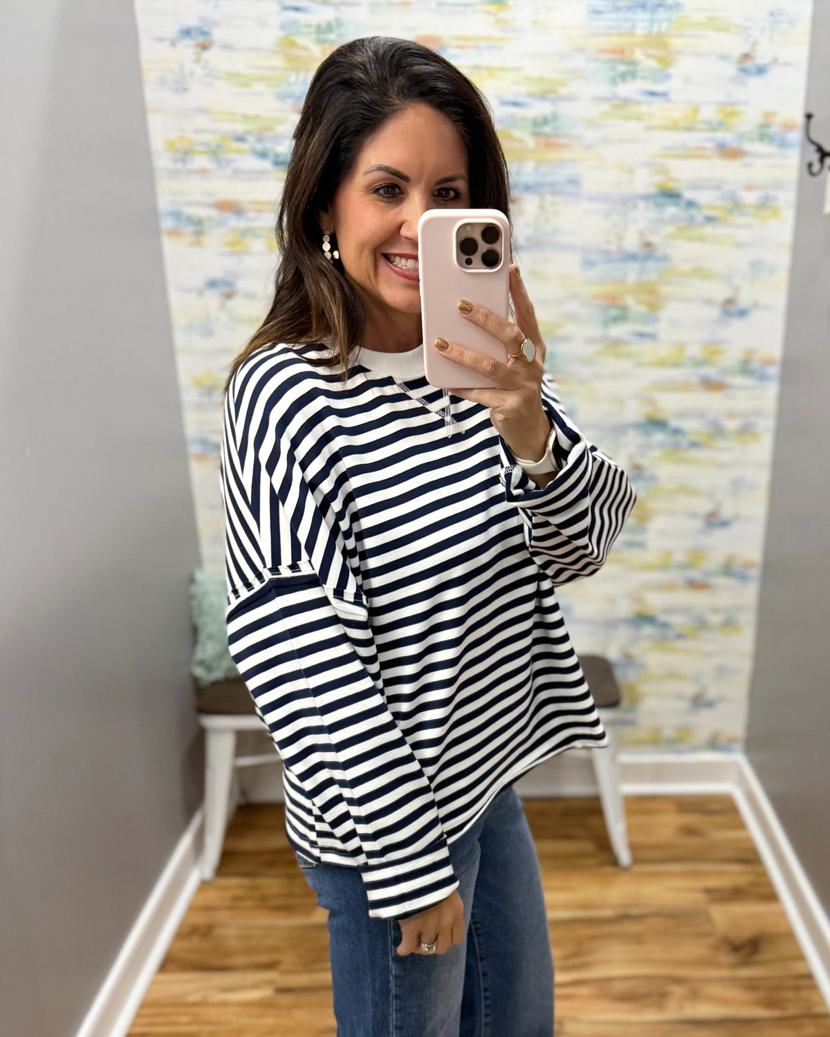 A woman takes a mirror selfie wearing a navy and white horizontally striped long-sleeve top, blue jeans, and a white undershirt in a fitting room with a patterned wallpaper background.