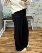 A person wearing black wide-leg pants and a cream sweater, standing in a room with wood floors and a bench in the background.