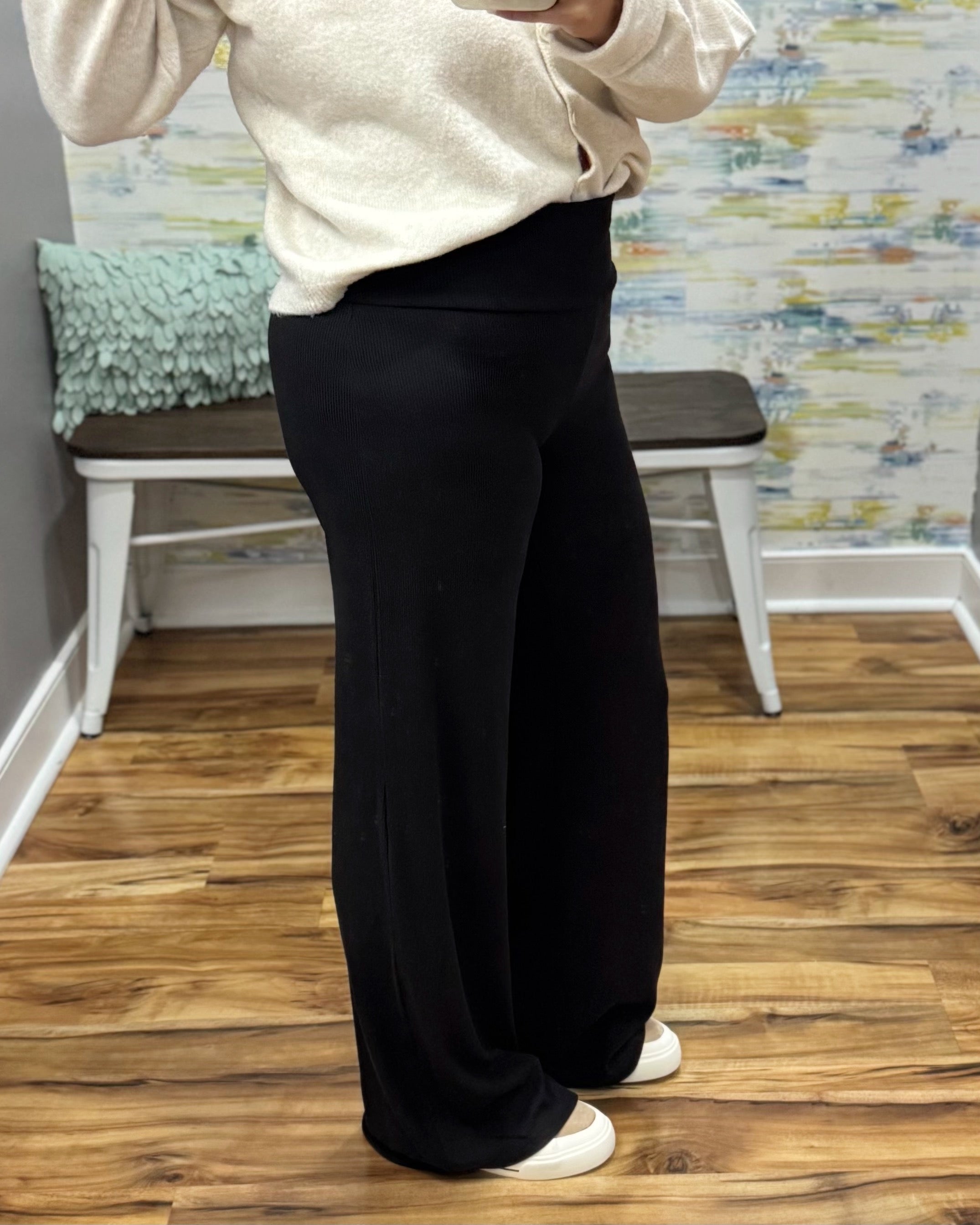 A person wearing black wide-leg pants and a cream sweater, standing in a room with wood floors and a bench in the background.