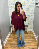 A woman takes a mirror selfie in a fitting room wearing a loose-fitting burgundy top and light blue flare jeans.