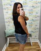 Woman taking a mirror selfie wearing a black sleeveless top and denim shorts in front of a colorful wall.