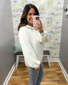 A woman takes a mirror selfie wearing a light heather gray hoodie and blue jeans in a fitting room with patterned wallpaper and wood floors.