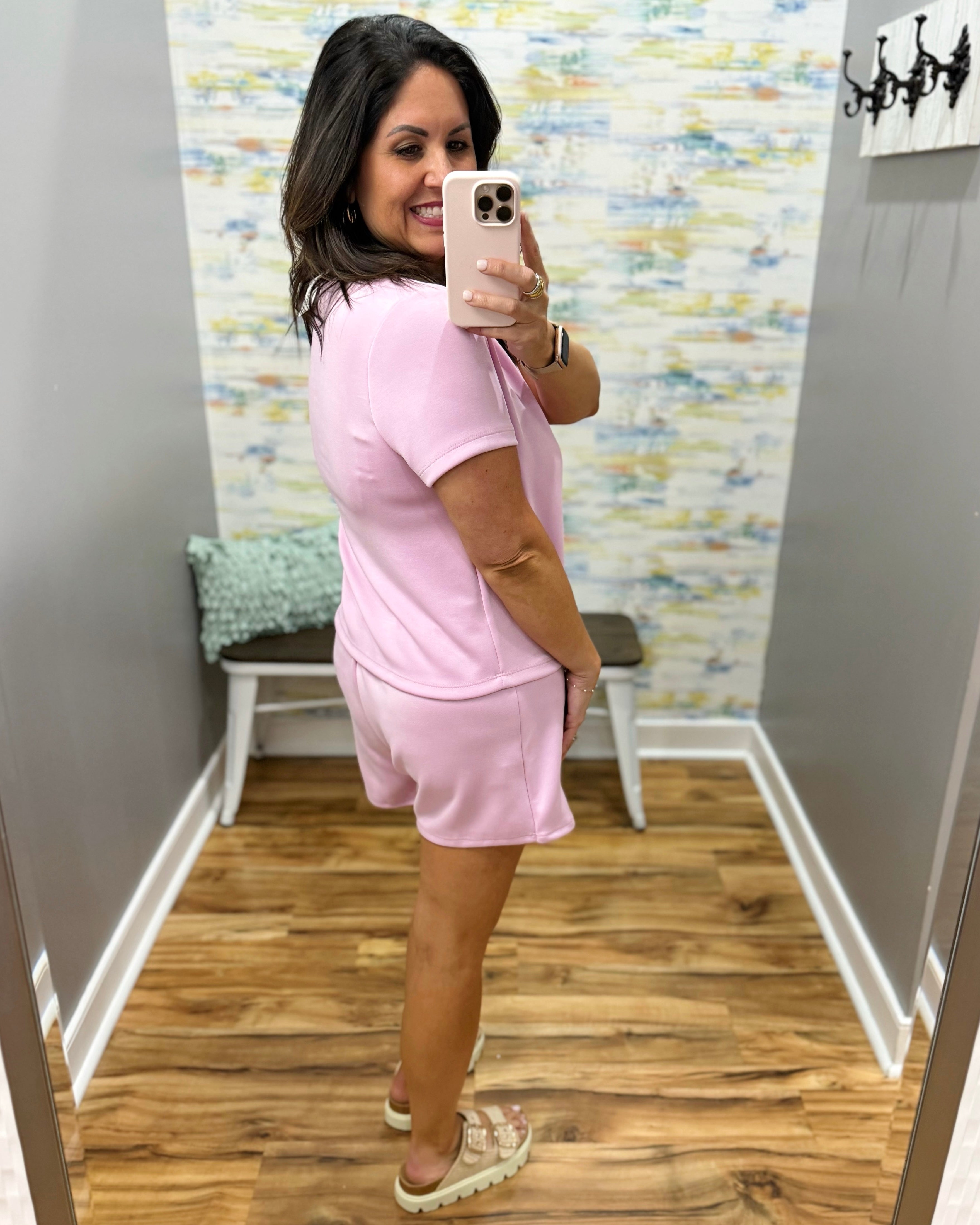 Woman taking a mirror selfie wearing a pink outfit in a room with patterned wallpaper.