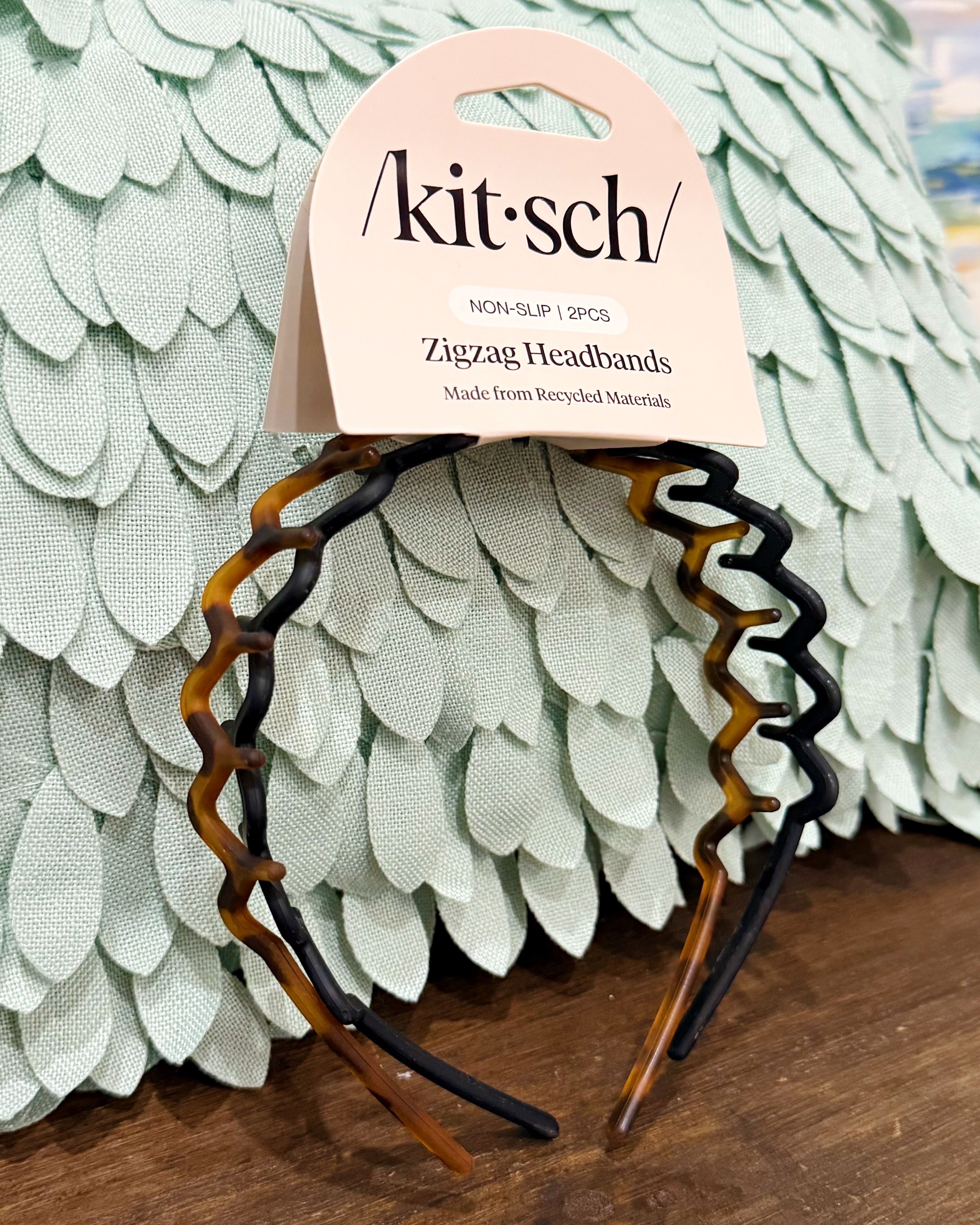 A pair of Kit·sch zigzag headbands in a tortoiseshell pattern, attached to a light pink paper header card. The headbands are displayed against a decorative, light green textured background on a wooden surface.