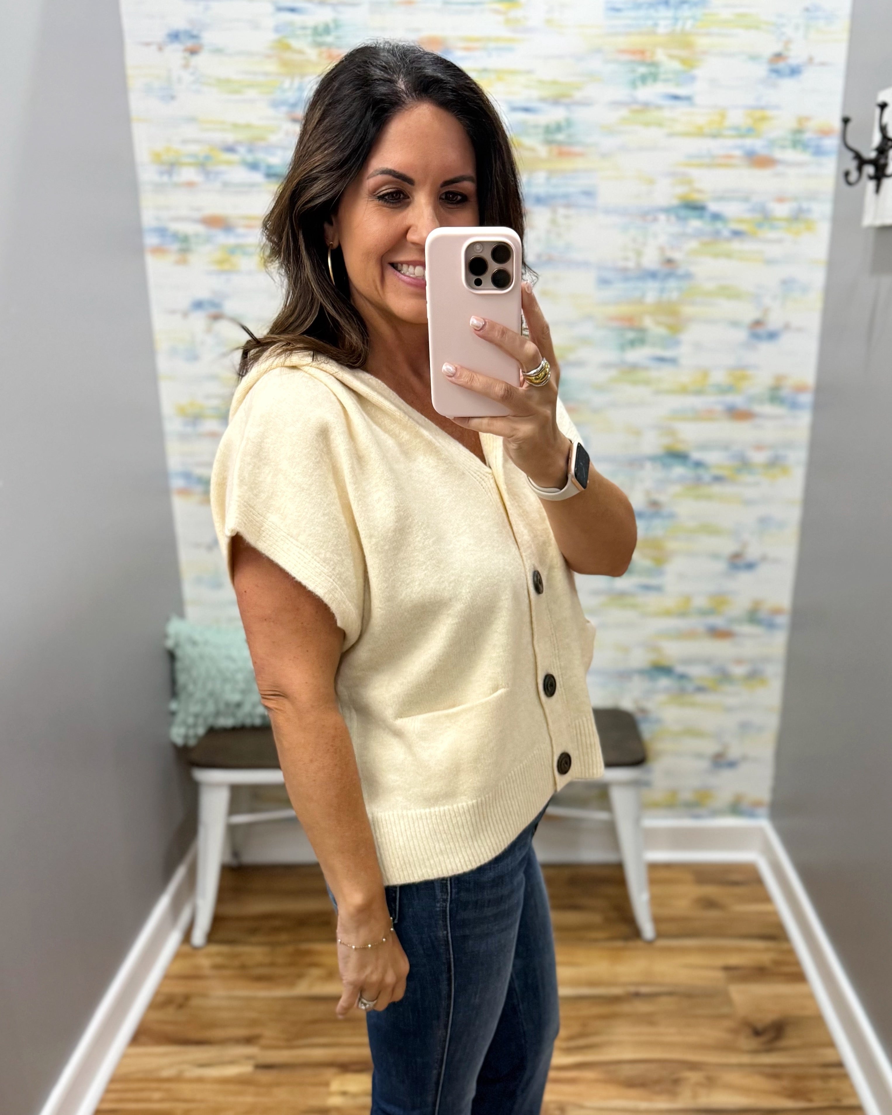 A woman takes a mirror selfie in a fitting room wearing a cream-colored short-sleeve knit cardigan with black buttons and dark blue jeans.