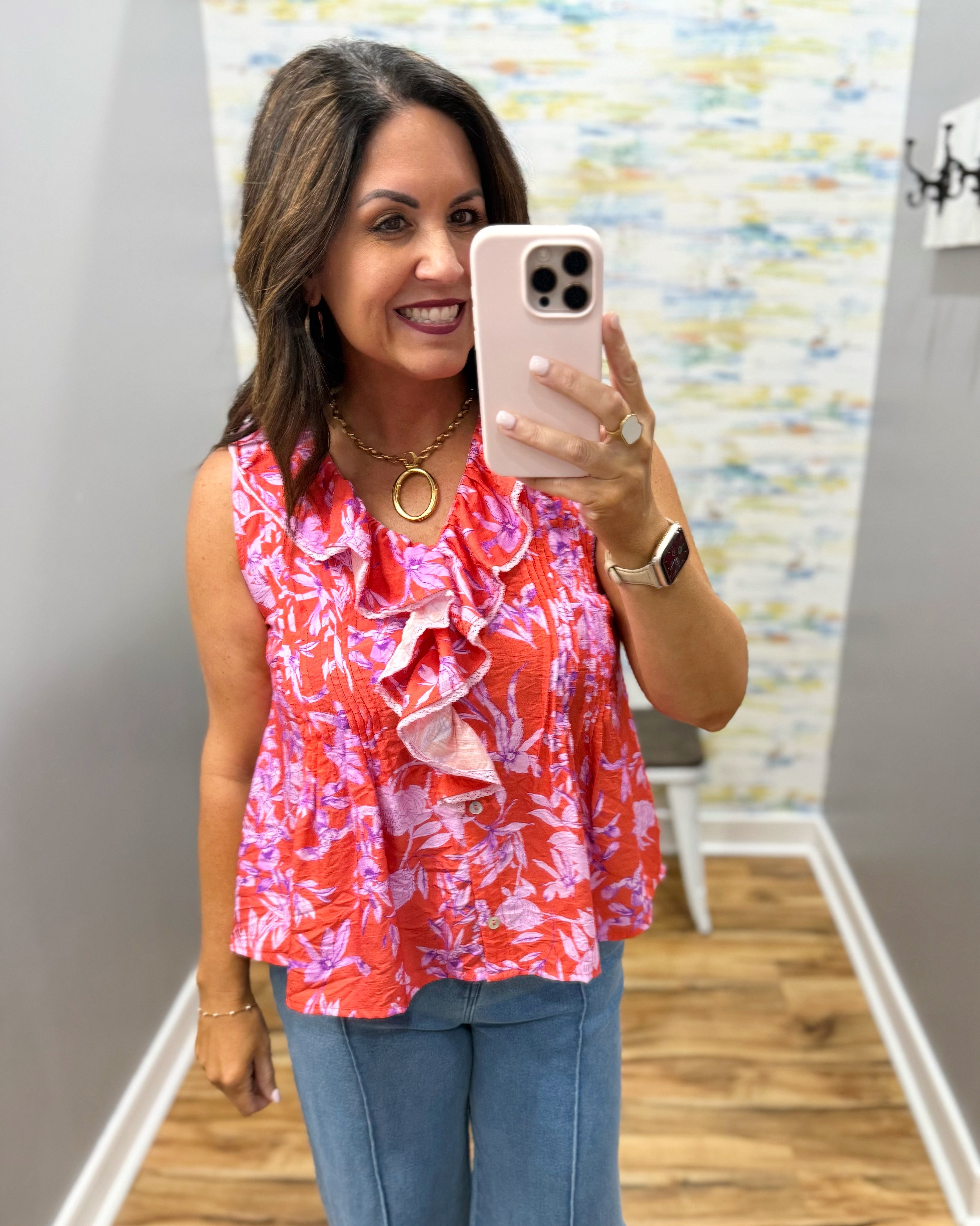 Woman taking a mirror selfie wearing a colorful blouse and jeans in a room with a patterned wall.