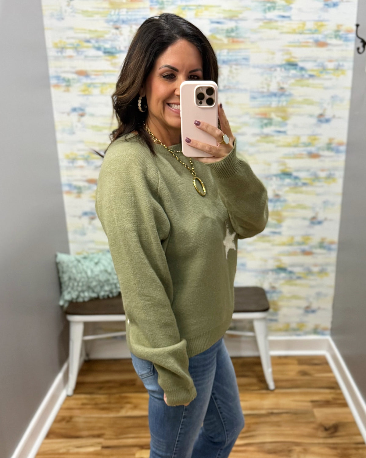 A woman takes a mirror selfie wearing an olive green knit sweater with a white star detail on the front, paired with blue jeans and gold jewelry, in a home setting with a patterned wallpaper background.