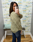 A woman takes a mirror selfie wearing a sage green and beige plaid long-sleeve blouse with dark blue jeans, standing in front of a patterned wallpapered wall.