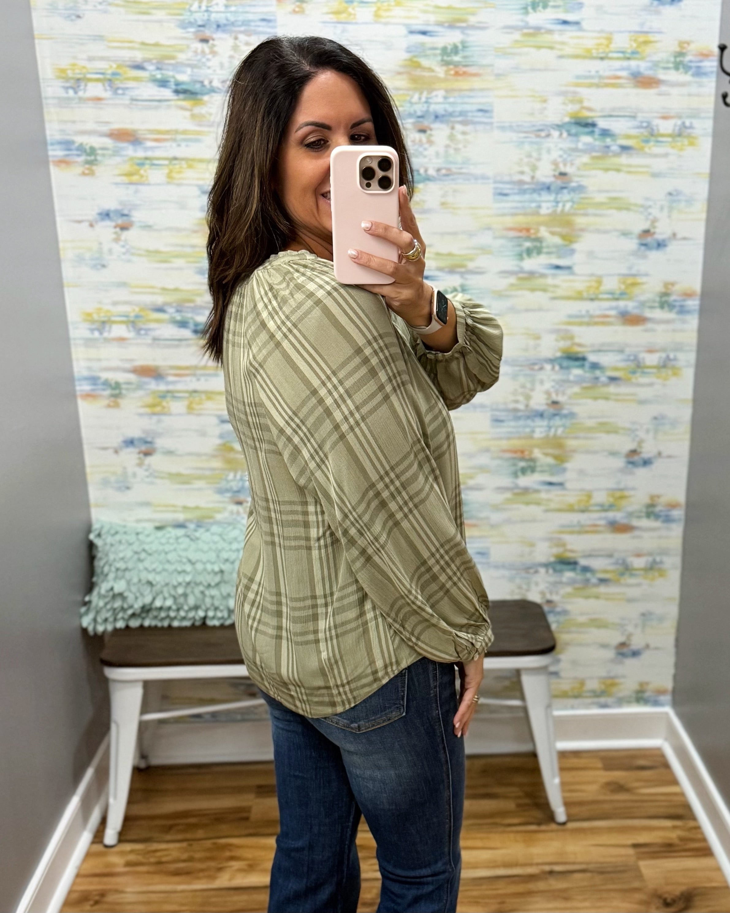 A woman takes a mirror selfie wearing a sage green and beige plaid long-sleeve blouse with dark blue jeans, standing in front of a patterned wallpapered wall.