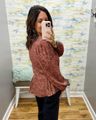 A woman takes a mirror selfie wearing a rust-colored long-sleeve blouse with a small floral pattern and dark pants in a home setting with patterned wallpaper.