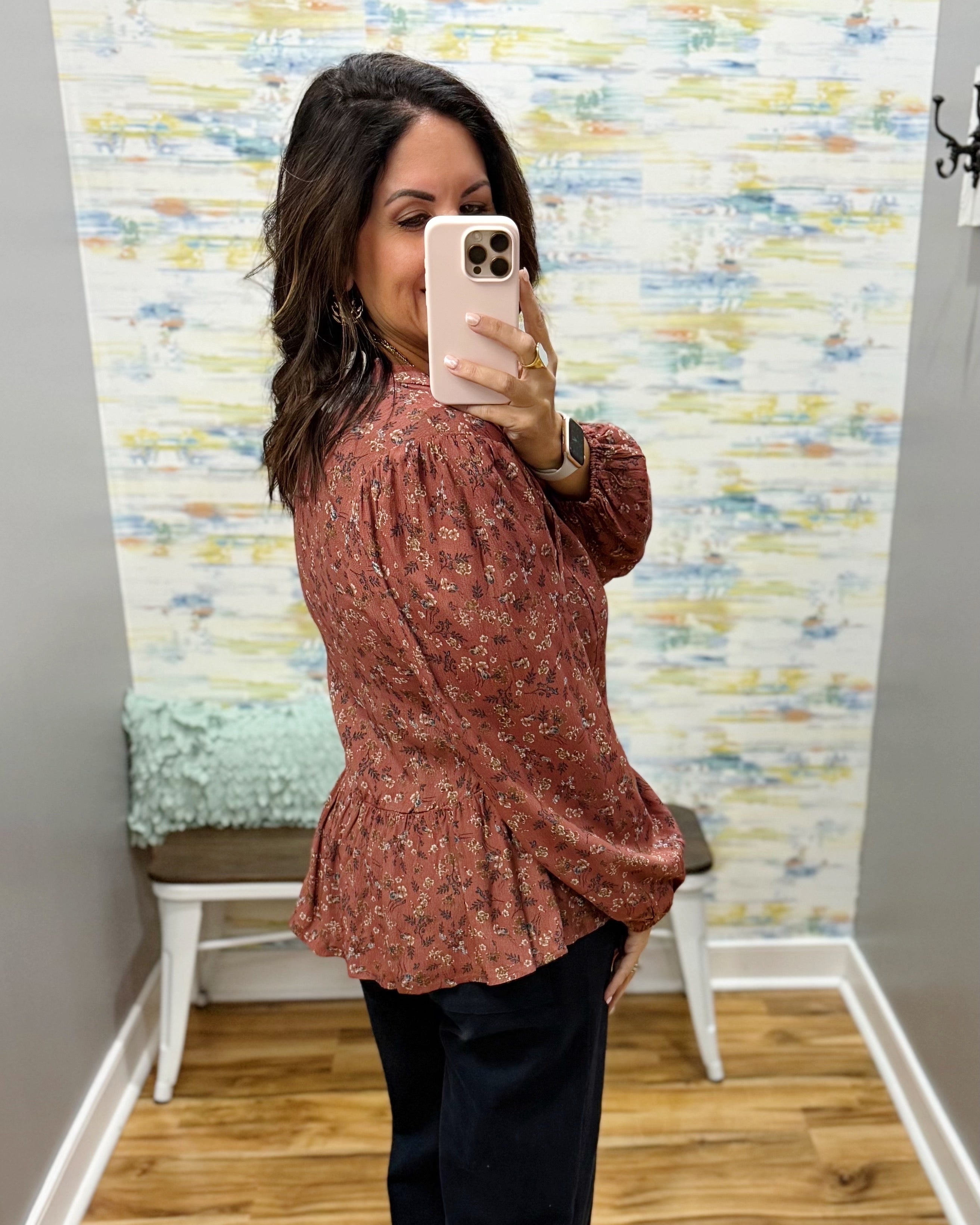 A woman takes a mirror selfie wearing a rust-colored long-sleeve blouse with a small floral pattern and dark pants in a home setting with patterned wallpaper.