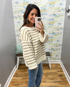 A woman takes a mirror selfie wearing a beige and white striped knit sweater, blue jeans, and a light pink phone in a home setting with wood floors and a patterned wallpaper background.