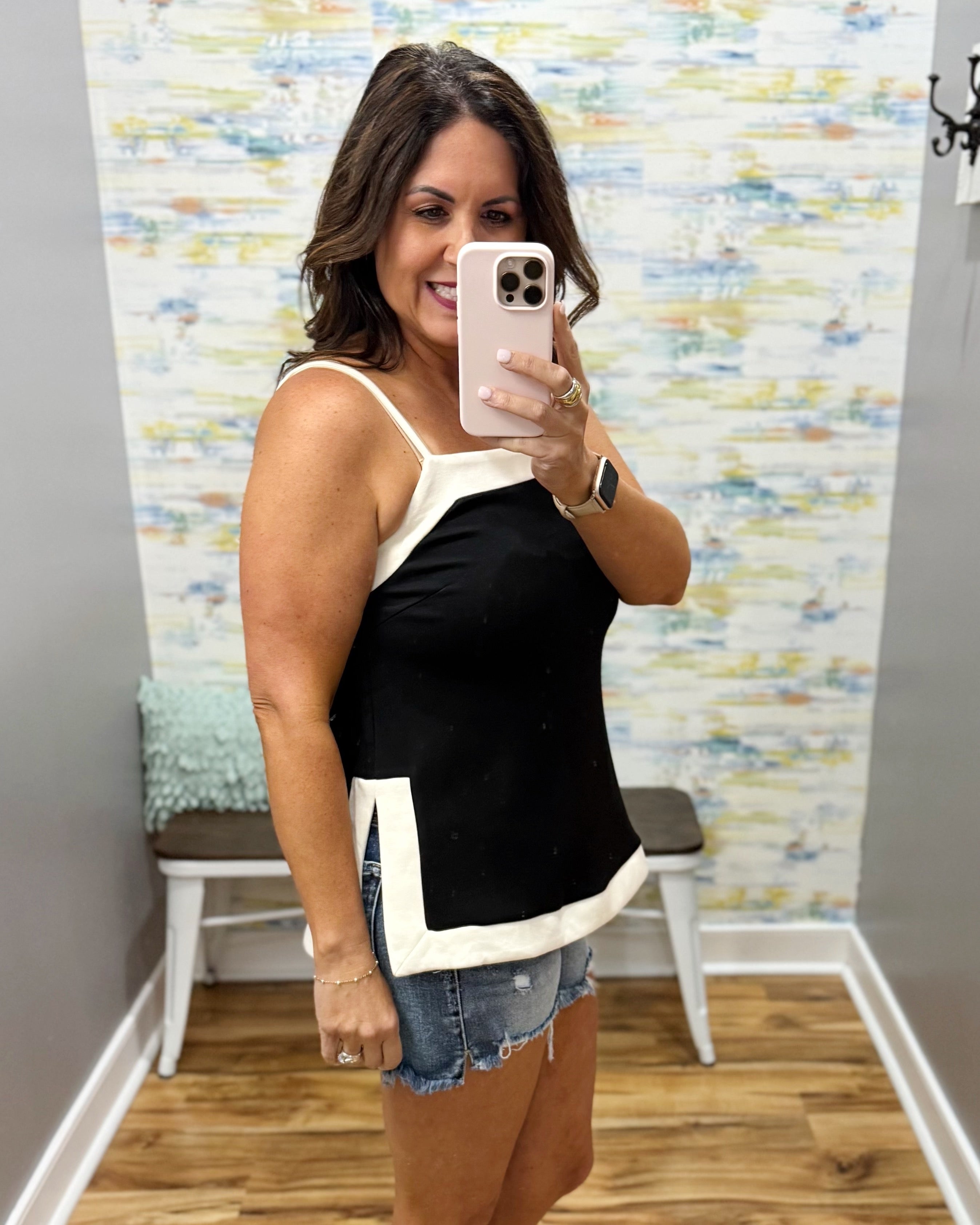 Woman taking a mirror selfie wearing a black and white tank top and denim shorts in a room with a colorful wall.