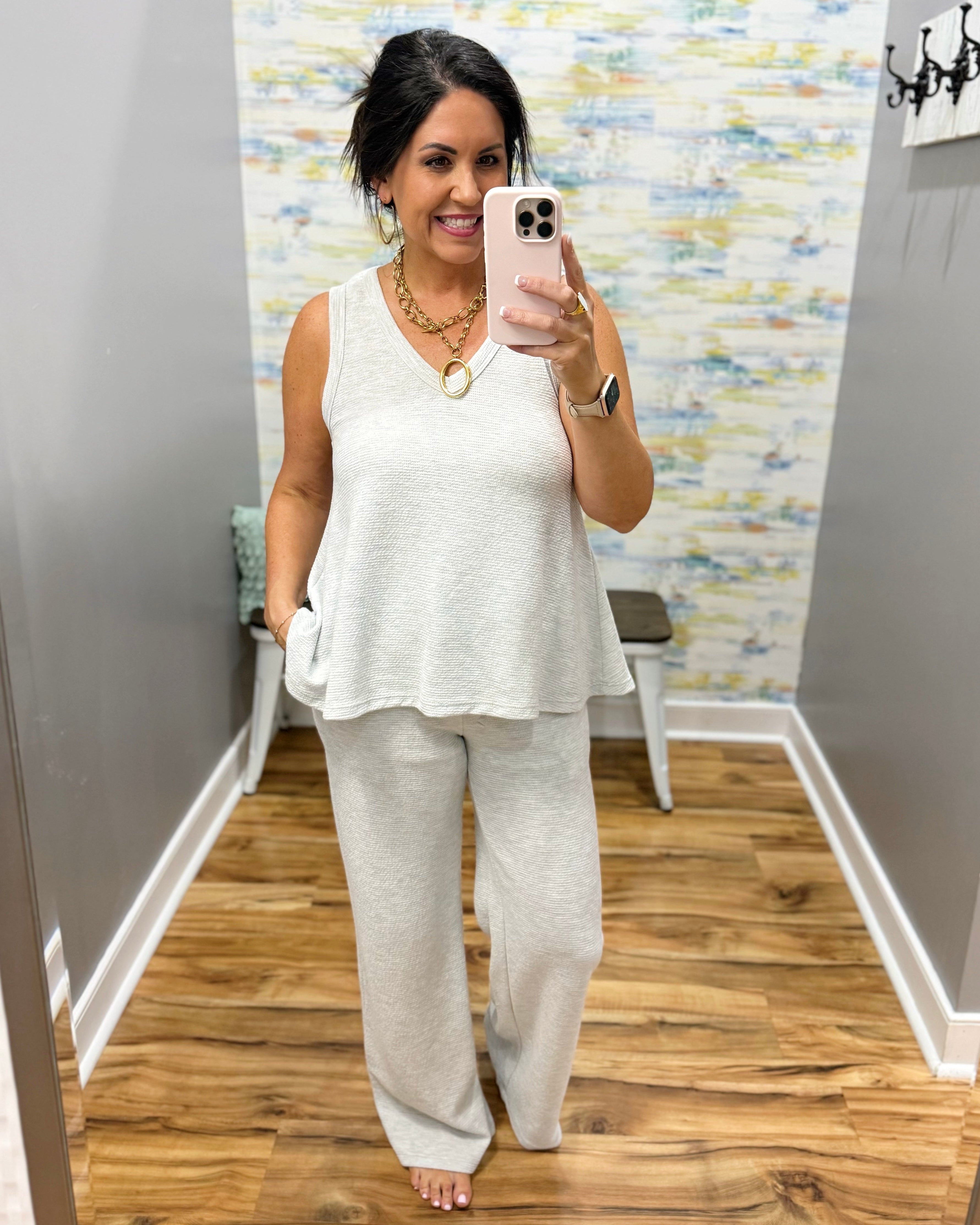 Woman taking a mirror selfie wearing a white sleeveless top and light gray pants in a room with a patterned wall.