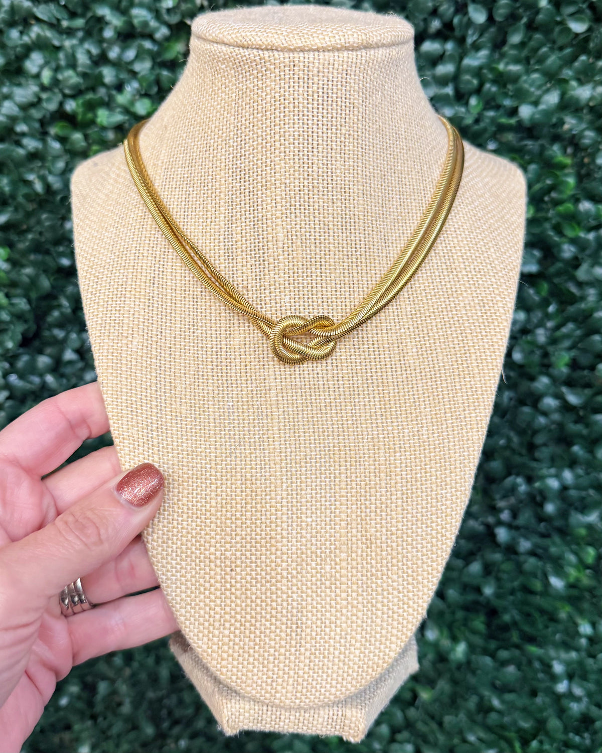 A gold-toned double-strand necklace with a knotted center detail, displayed on a beige fabric jewelry bust. A hand with glittery nail polish is holding the left side of the bust, and a green leafy background is visible behind it.
