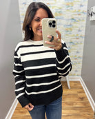 A woman taking a mirror selfie wearing a black and white horizontally striped knit sweater, blue jeans, and holding a light-colored phone.