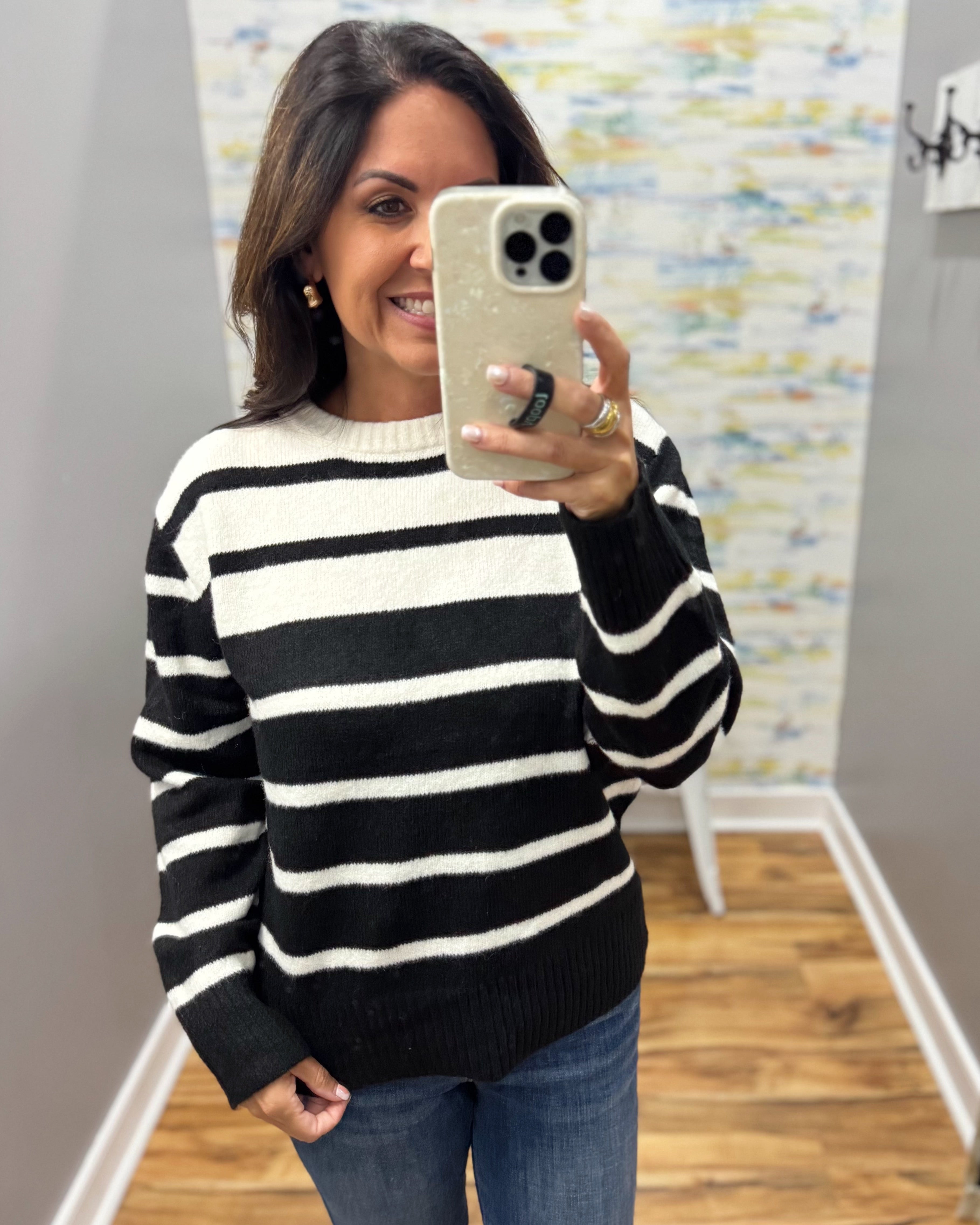 A woman taking a mirror selfie wearing a black and white horizontally striped knit sweater, blue jeans, and holding a light-colored phone.