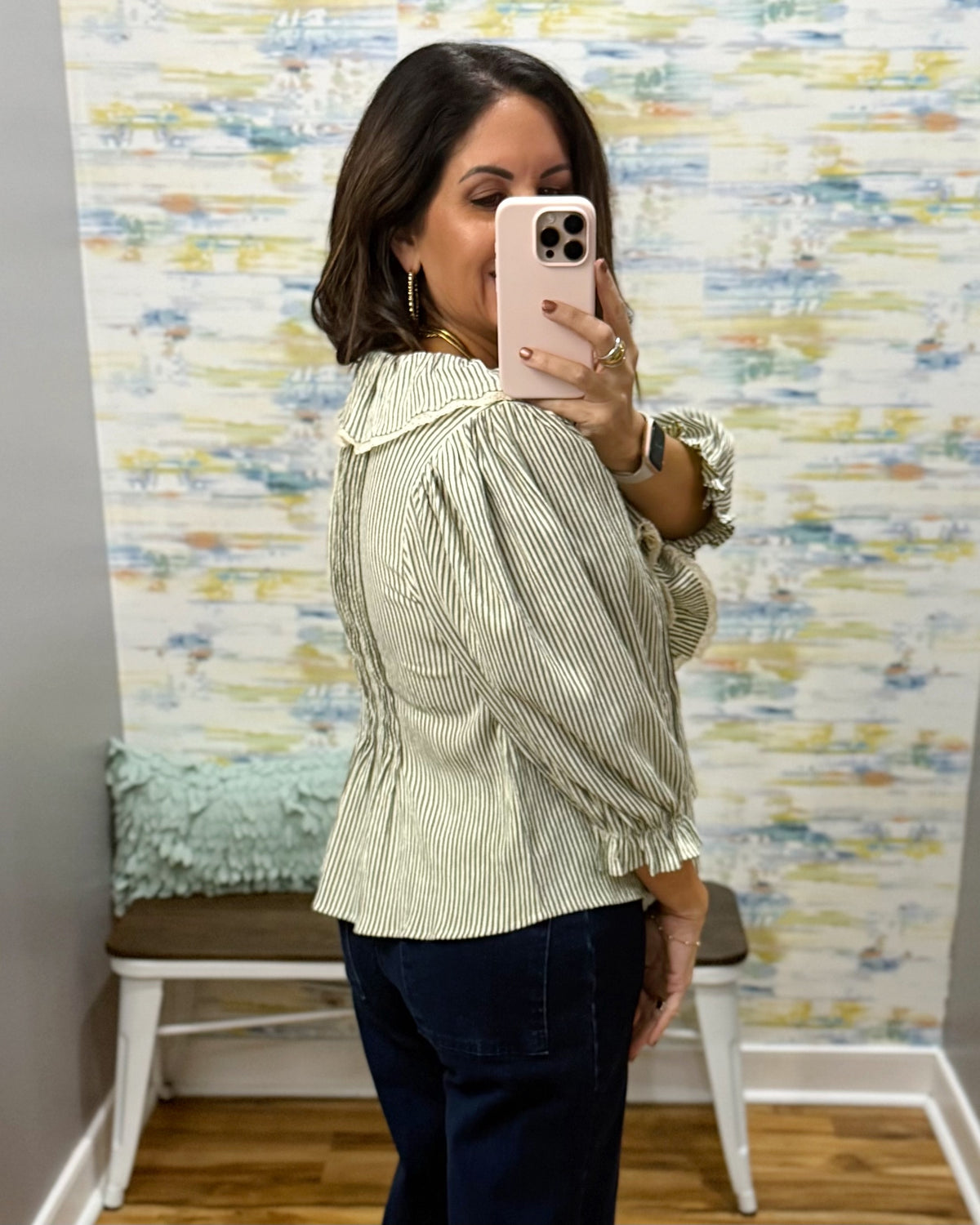 A woman takes a mirror selfie wearing a light sage green and white vertically striped blouse with ruffled details, paired with dark blue pants, in front of a patterned wallpaper background.