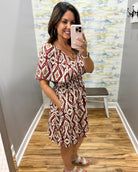 A woman takes a mirror selfie in a boutique wearing a short-sleeve V-neck dress with a red, beige, and white geometric diamond pattern. The background shows a gray wall, a colorful abstract wallpaper, a wooden floor, and a small bench.